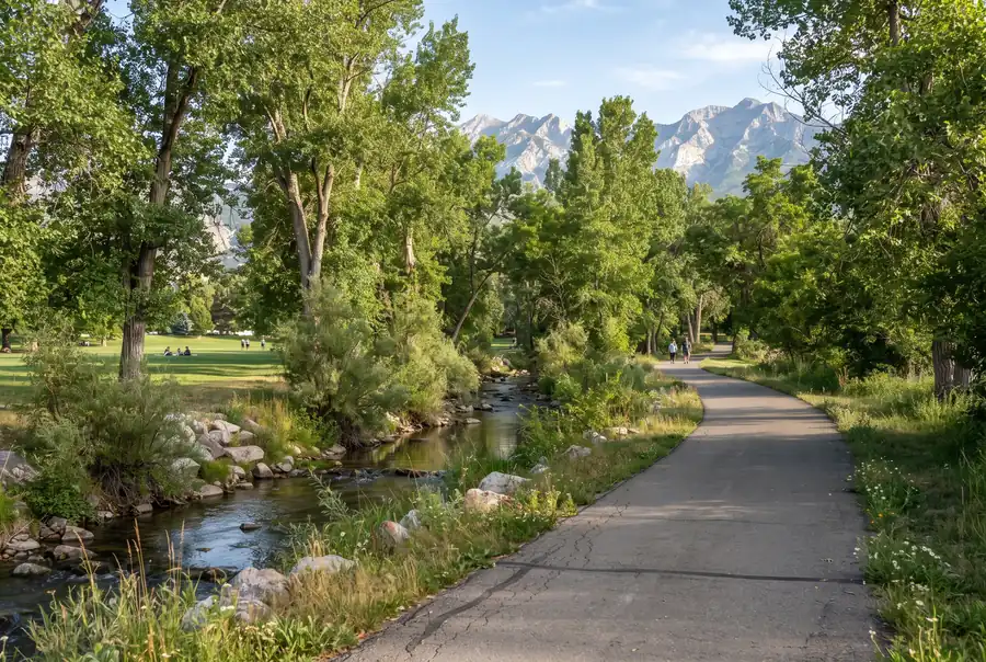 Tree-lined park path with a stream and mountain backdrop in Murray, Utah
